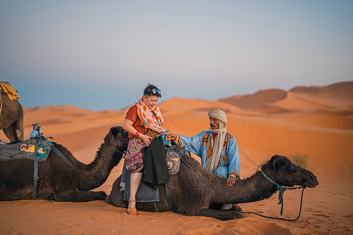 Asian Chinese female tourists getting up riding dromedary camel train crossing Sahara Desert Morocco