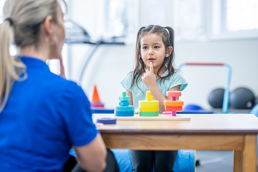 A female therapist sits with a sweet little girl as a table as they work on her speech together.  The Therapist is dressed professionally and is pointing to her mouth to help show the little girl how to form her words.