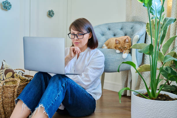istock Middle aged woman using laptop, with sleeping cat lying on armchair at home 1464016366