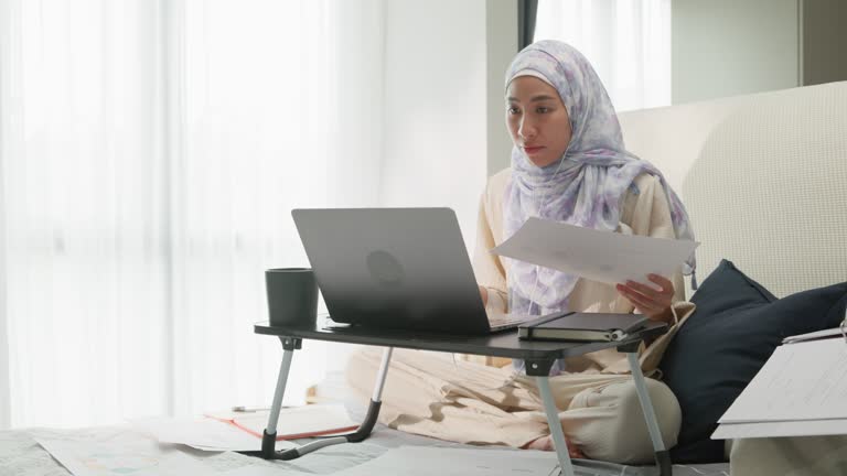 Happy young muslim woman sit on bed with computer laptop and coffee cup on folding laptop table, portable holder stand hold paperwork video online meeting in bedroom at home. Work from home.