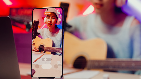 Close-up Happy Asian musician girl influencer wear headphone play guitar record phone podcast online live stream sing song with follower audient at studio. Female podcaster, Audio podcast concept.