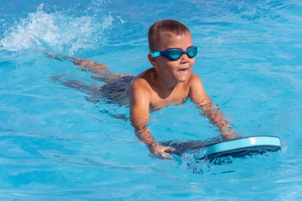 Photo of Happy boy child in swimming goggles with float foam board tool swim in pool in sunny day. Safe pool training, summer holiday, family vacation, travel, water sport concept