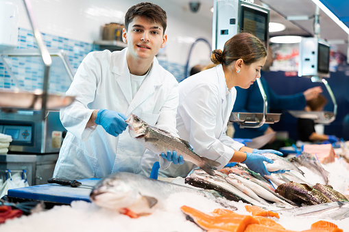 Positive young salesman demonstrating seabass behind counter in fish store with large assortment