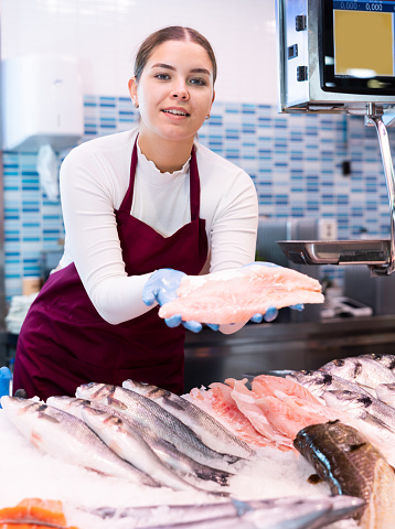 Positive young saleswoman in vinous apron demonstrating fish fillet behind counter in fish store