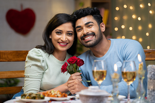 Happy young couple holding red rose by looking at camera during candlelight dinner at home - concept of valentines day, togetherness and dating.