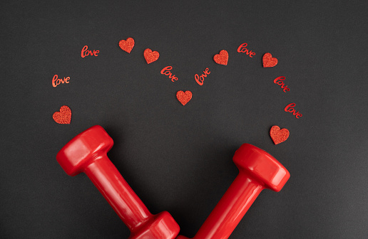 Gym dumbbells and red decorations in shape of a heart. Love gift for Valentine's Day, marriage proposal engagement, anniversary or wedding. Fitness workout and sport training flat lay composition with copy space.
