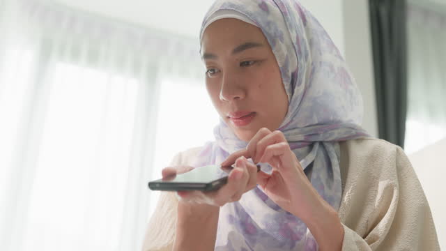 Closeup of happy young muslim woman sit on bed with computer laptop and use smartphone giving voice commands to virtual assistant in bedroom at home. Work from home.