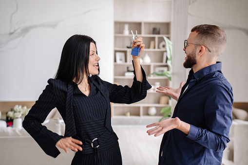 Young caucasian couple celebrating new apartment. Excited Italian woman holds keys of flat dancing looks at husband. Handsome beardy man surprised after purchasing home. Mortgage