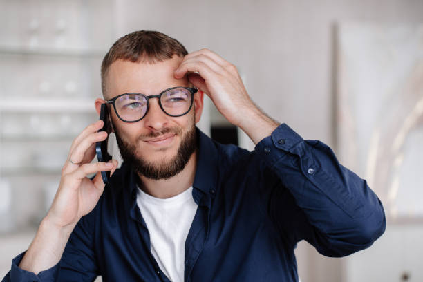 un jeune homme caucasien barbu bouleversé dans des lunettes parle par téléphone prend une décision. l’entrepreneur américain discute avec son partenaire hésite. un italien réfléchi en décontracté parle à la maison par smartphone. - facial expression businessman bad news surprise photos et images de collection