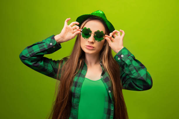 Haughty stylish young woman wearing party green hat, clover shaped glasses, checkered plaid shirt and looking aside isolated on colored green background. St Patricks Day celebration concept. Haughty stylish young woman wearing party green hat, clover shaped glasses, checkered plaid shirt and looking aside isolated on colorful green background. St Patricks Day or March 17 celebration concept. monochrome clothing stock pictures, royalty-free photos & images