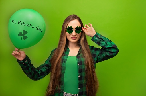 Smiling girl in green checkered plaid shirt with clover shaped glasses holding green air balloon with text words St Patrick's Day and shamrock on colorful green background. March 17 or St Patricks Day celebration concept.