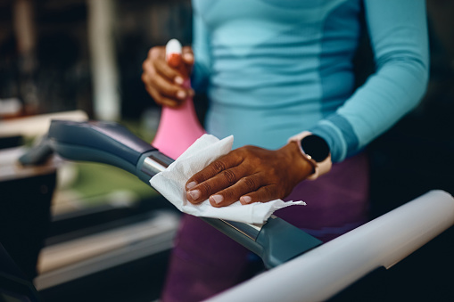 Close up of African American athletic woman spraying treadmill with disinfectant before exercising in gym