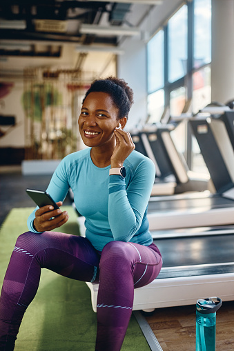 Happy African American athletic woman using mobile phone while listening music over earphones in a gym and looking at camera.