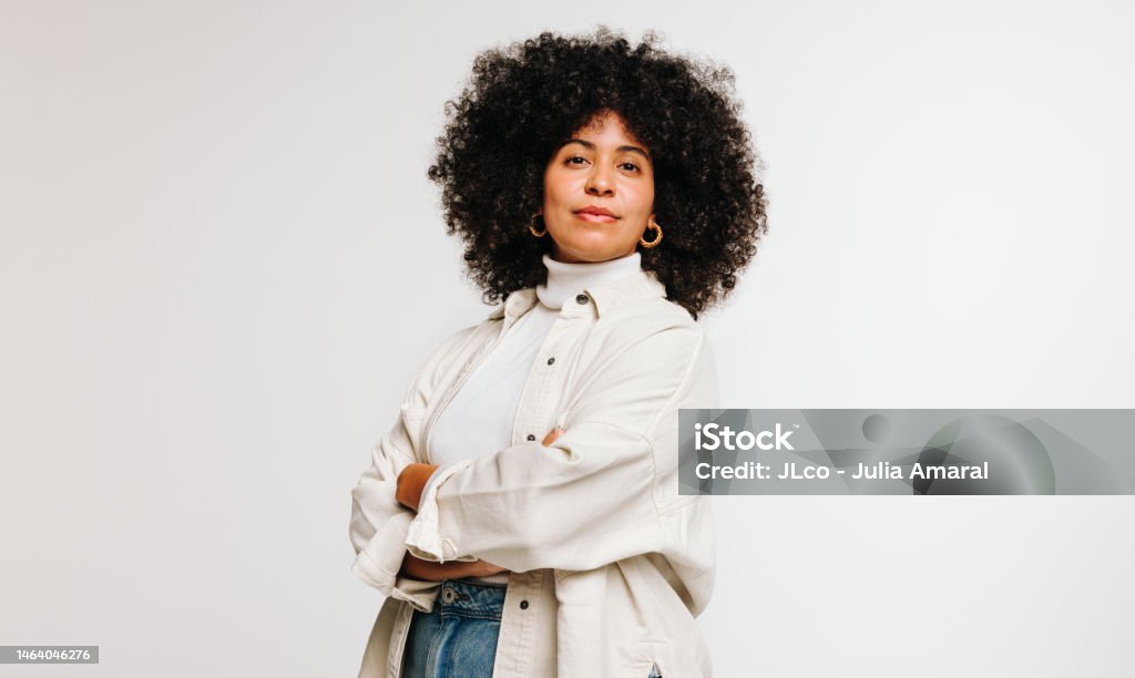 Self-confident woman with an Afro hairstyle standing in a studio Self-confident woman with an Afro hairstyle looking at the camera while standing against a studio background. Attractive young woman of colour wearing her curly hair with pride. Women Stock Photo