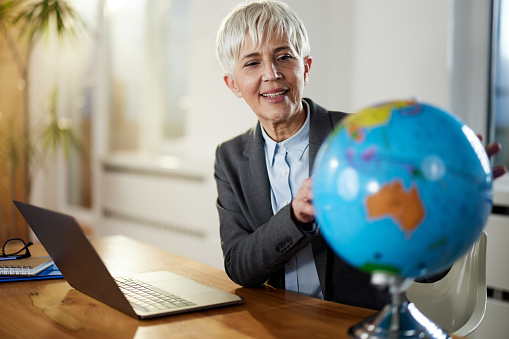 Happy mature female entrepreneur analyzing the globe during online meeting at home office.
