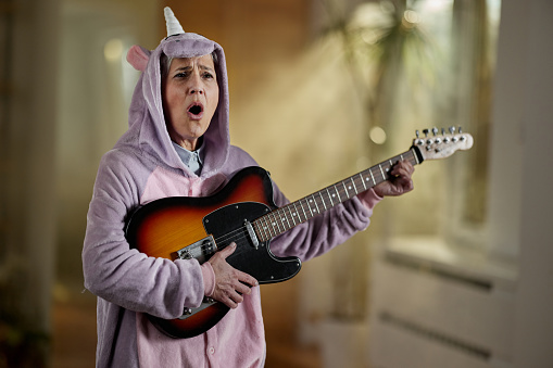 Mature woman in unicorn costume having fun while singing and playing a guitar at home.