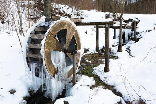 In a creek a wooden water wheel in the winter is covered with thick icicles in a snowy landscape.