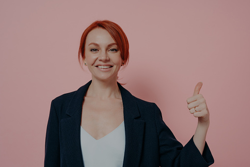 All good. Isolated studio shot of young successful cheerful red-haired business woman showing thumb up hand gesture, demonstrating acceptance, saying i like it and smiling at camera, isolated on pink