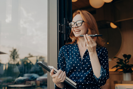 Positive beautiful woman with red hair holds phone talks on speakerphone with friend makes voice recognition or request uses internet services poses near cafeteria wears spectacles polka dot dress