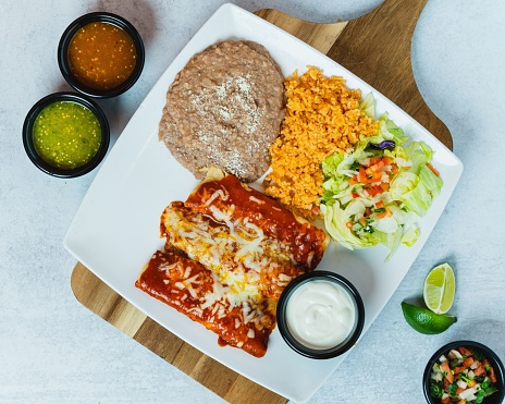 A top view of red enchiladas on a wooden board with refried beans, rice, salsa, and lime