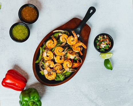 A top view of shrimp fajitas in a pan on a wooden board with salsa, pico de gallo, and peppers
