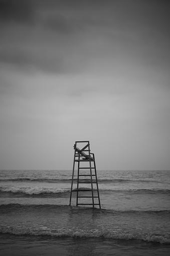 A vertical grayscale shot of a lifeguard chair in the shallow sea under dark clouds