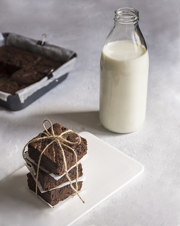 We see a stack of three brownies wrapped and served on a white plate. in the background, we can see a bottle of milk.