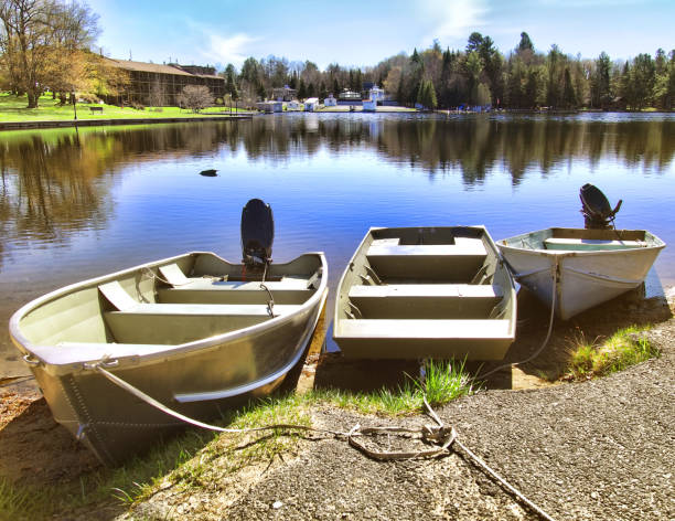 The quiet harbor scene in Forland, a village on the island of Sotra, Norway. Two boats—one recreational and one fishing vessel—are positioned on land near a wooden dock, adjacent to a red and white boathouse with a sloped roof. The calm water in the foreground reflects the dock and surrounding structures. The backdrop features rocky hills covered in moss, shrubs, and evergreen trees under an overcast sky. This image highlights the intersection of natural beauty and coastal utility in a Nordic maritime setting.