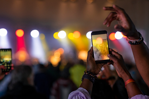 A point of view shot of an unrecognisable woman at a festival taking a picture on her smartphone of a live music festival. Everyone in the crowd is dancing together and having a good time. They are happy and sharing positive energy while watching the live show.