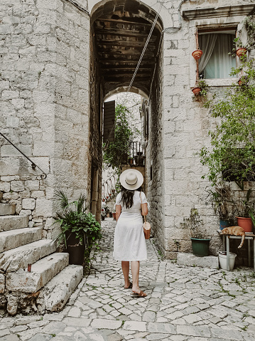 Rear view of young woman wearing white dress and sun hat holding handbag. Lifestyle and fashion full length image of female tourist in old town.