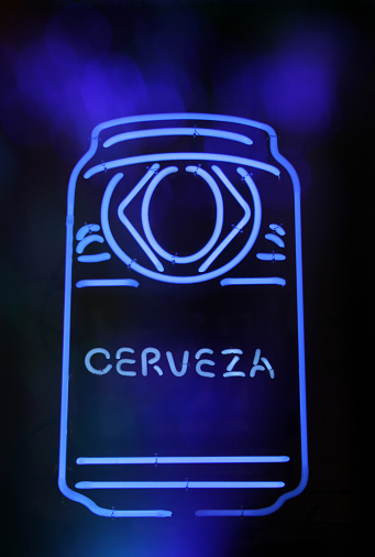 A vertical shot of a blue neon beer can sign with a Spanish text Cerveza (beer) on a dark background