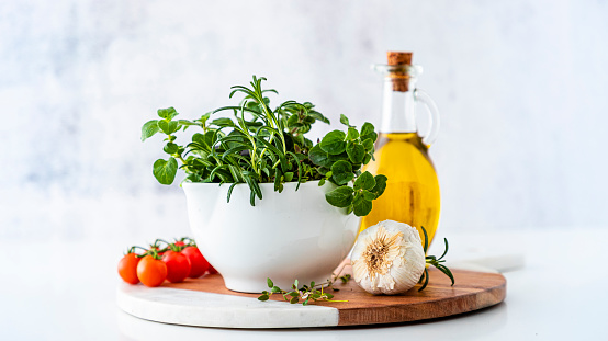 Italian ingredients: Front view of a mortar full of fresh aromatic herbs such as rosemary, thyme and oregano surrounded by a garlic bulb, some cherry tomatoes and an olive oil bottle on a white defocused countertop. Objects are at the center of the image.