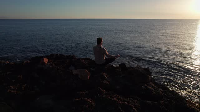 Focused man practicing meditation on a rocky beach summer morning. Yoga man sitting in lotus position near ocean waves. Attractive sportsman doing relaxing asana outdoors. Self care concept.