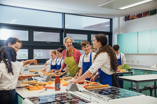 A side view of a teacher supervising her students as they make pizza in a cooking class in the Northeast of England. They are wearing aprons to keep their school uniforms clean. She is helping them with their choices and with their safety using the equipment.