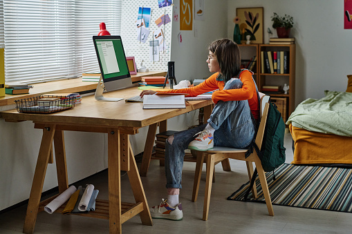 Teenage girl sitting on table in front of computer monitor with books and studying online at home