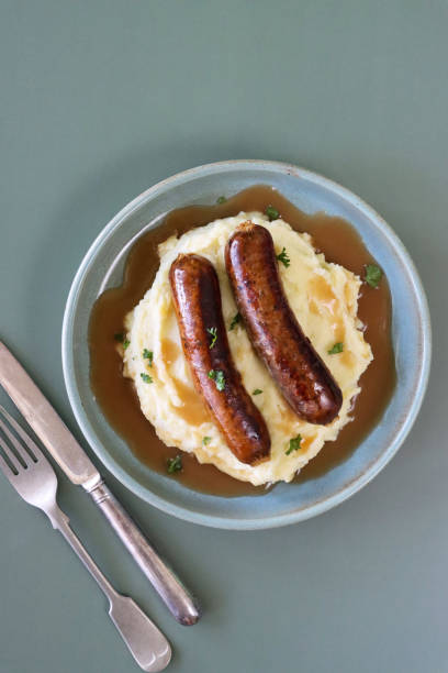 Stock photo showing elevated view of homemade bangers and mash with rich onion gravy covering sausages and mashed potato. imagen del plato de bangers y puré tradicionales británicos, montículo de puré de papas cubierto por dos salchichas de cerdo cubiertas con salsa de cebolla, cuchillo y tenedor de metal, comida reconfortante abundante, fondo verde, vista elevada