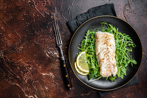 Grilled cod fish fillet served with green salad in a plate. Dark background. Top view. Copy space.