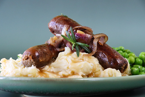 Stock photo showing close-up view of homemade bangers and mash with rich onion gravy covering sausages and mashed potato served with garden peas.
