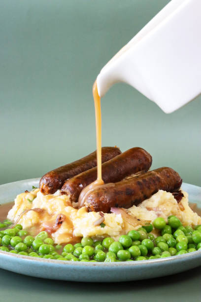 immagine del sugo che viene versato dalla barca di sugo sul piatto dei tradizionali bangers e purè britannici, tumulo di purè di patate sormontato da tre salsicce di maiale su un letto di piselli verdi da giardino, abbondante cibo di conforto, sfondo ver - sausage food mash grilled foto e immagini stock