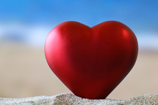 Stock photo showing close-up view of a red heart-shaped model on sand pile on a sunny beach with the sea and clear blue sky in the background. Holiday vacation romance concept.