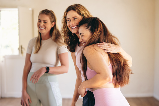 Two women greeting each other with a hug in a yoga studio. Happy female friends attending a yoga class in a community wellness centre. Women of different ages starting a fitness session.