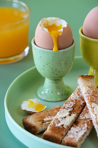 Stock photo showing close-up view of green and yellow crockery of tea plate and egg cups with buttered, white toast soldiers and two soft-boiled eggs one topped showing runny yellow yolk.