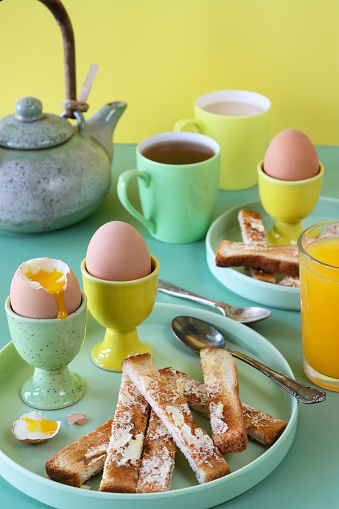 Stock photo showing close-up view of green and yellow crockery of tea plates and egg cups with buttered, white toast soldiers and soft-boiled eggs one topped showing runny yellow yolk.