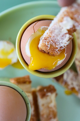 Stock photo showing close-up, elevated view of green and yellow crockery of tea plate and egg cups with buttered, white toast soldiers and two soft-boiled eggs one topped showing runny yellow yolk.