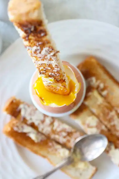 Stock photo showing close-up, elevated view of white crockery of tea plate and egg cup with white toast soldiers and topped soft-boiled egg showing runny yellow yolk.