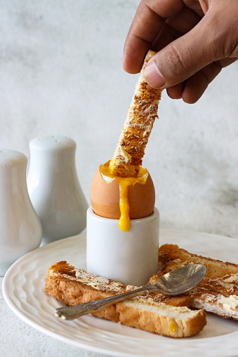 Stock photo showing close-up view of white crockery of tea plate and egg cup with white toast soldiers and topped soft-boiled egg showing runny yellow yolk.