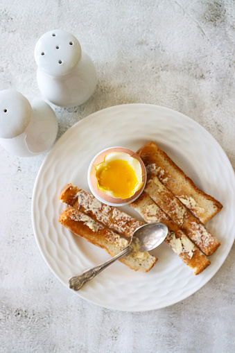 Stock photo showing elevated view of white plate containing eggcup with boiled dippy egg and buttered, white toast soldiers for breakfast.