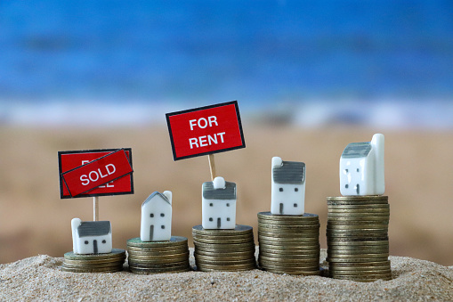 Stock photo showing close-up view of 'For Sale' sign with diagonal 'Sold' banner across text besides row of model coastal houses stood on stacks of coins on a sunny beach with the sea and clear blue sky in the background. Real estate, vacation home and beach hut rental concept.