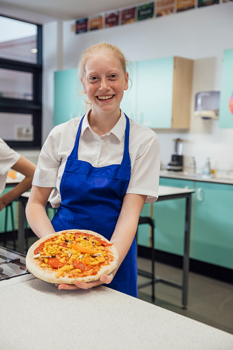 A close up of a young blonde female teenager holding out her pizza which she has chose the toppings for in her cooking class. She is smiling and looking into the camera in the school she goes to in the North East of England. She has made a pepperoni pizza with mushrooms and choose.
