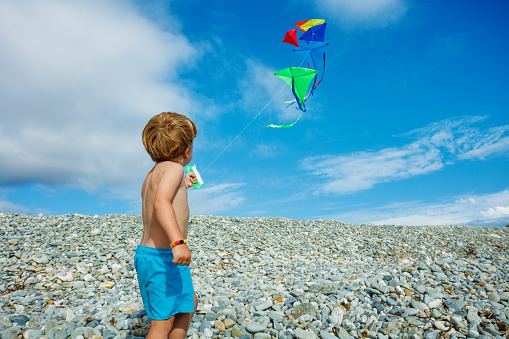 Little blond boy stand holding many kites in hand on pebble beach over blue sky